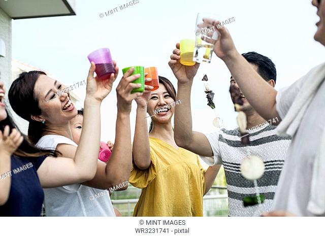Group of cheerful young Japanese men and women standing outdoors, toasting with plastic beakers