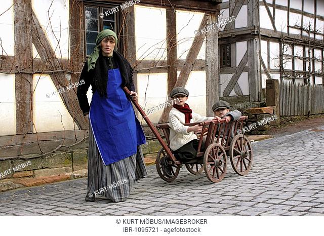 Woman wearing a costume from the 1920's pulling a hand cart with two children along a paved village street, public demonstration in Hessenpark, Neu-Anspach