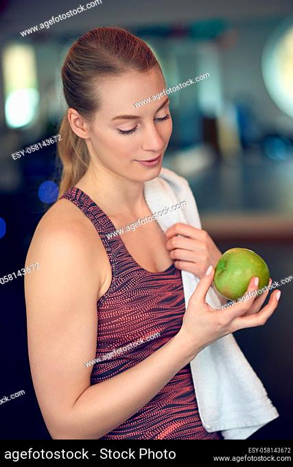 Fit sporty young woman contemplating a fresh green apple she is holding in her hand after working out in the gym in a concept of healthy diet and active...