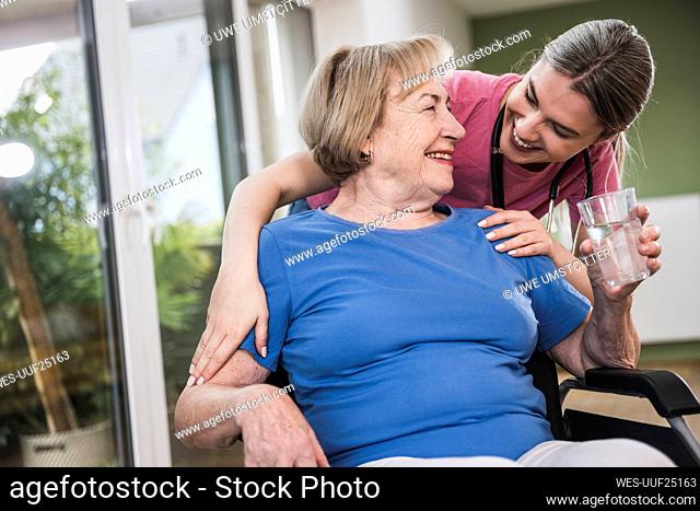 Smiling caregiver standing behind disabled woman in wheelchair at home