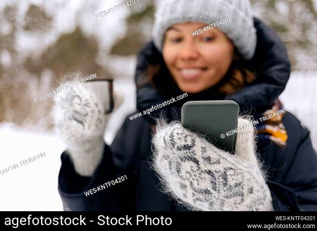 Woman with thermo mug taking selfie with smartphone in winter