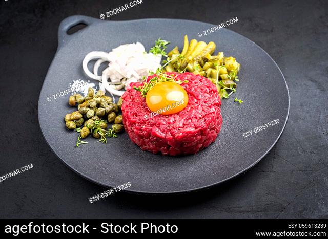 Gourmet tartar raw from beef fillet with yellow of the egg and gherkin with capers and onion rings as closeup on modern design plate