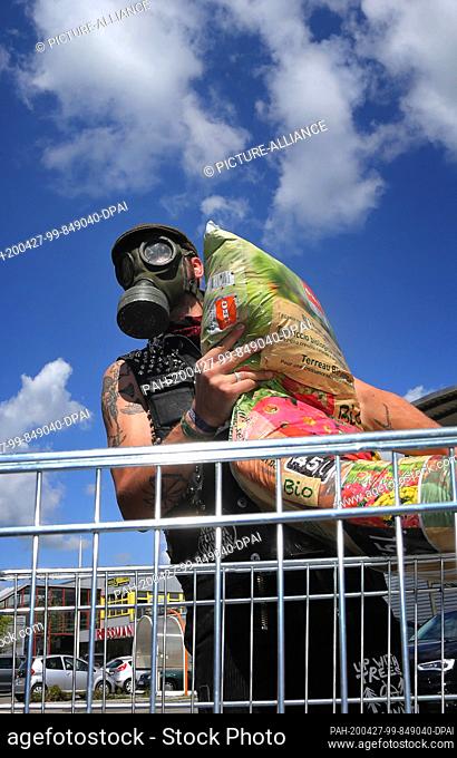 27 April 2020, Bavaria, Kaufbeuren: A young man wearing a gas mask loads the potting soil he has bought into his car in front of a hardware store