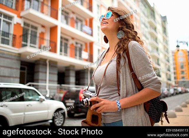 Traveling young woman in the european city. Girl with long curly hair in sunglasses holding old camera in her arms