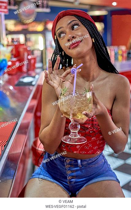 Young woman with braided hairstyle sitting on a bar, drinking a cocktail with a straw