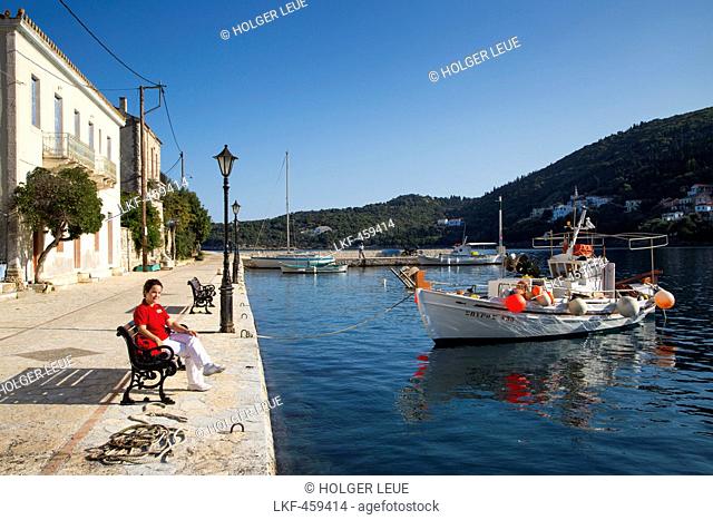 Woman sitting on a bench in the idyllic fishing village with fishing boat, Kioni, Ithaca, Ionian Islands, Greece