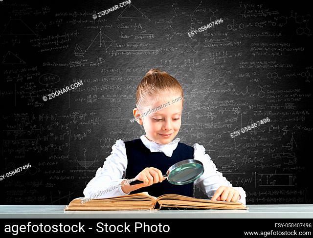 Little girl sitting at desk with magnifier and open book. Research and education in school. Happy schoolgirl in school wear carefully studying book