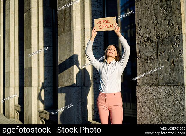 Smiling beautiful businesswoman holding open sign cardboard placard against building in financial district