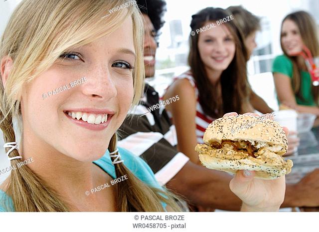 Portrait of a young woman holding a burger with her friends sitting in the background