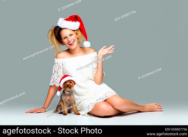 Beautiful happy girl in lace dress and red santa cap sitting with pretty yourkshire terrier in christmas hat. Studio shot over grey background