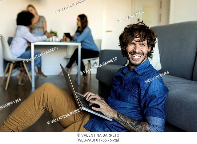 Laughing man sitting on floor using laptop with friends in background