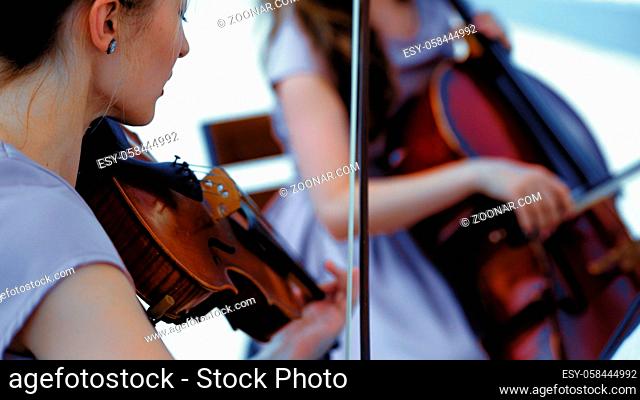 Musical Band Of Female Musicians Plays On Event Outdoor, Hands Holding A Violin And Bow, Close Up Cello on Blurred Background