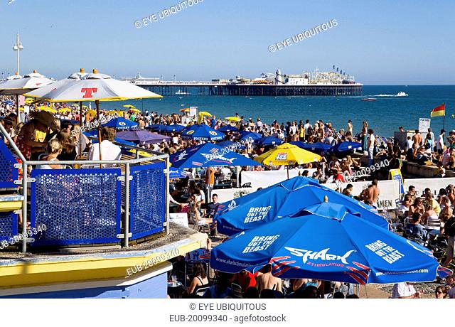 People sitting under sun shade umbrellas at tables on the promenade outside bars and restaurants with Brighton Pier and people on the shingle pebble beach...