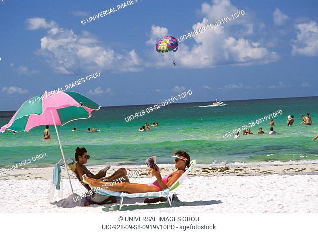 Beach Scene - Women Sunbathing On Lounge Chairs, People Swimming In Ocean, Boat And Paraglider In Distance