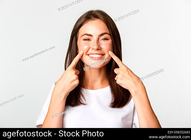 Close-up of attractive smiling young girl pointing fingers at face, poking cheeks, white background
