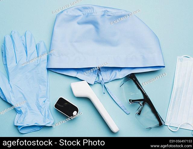 textile blue cap, disposable medical mask, pair of gloves and plastic glasses on a blue background, top view. Medic protective clothing
