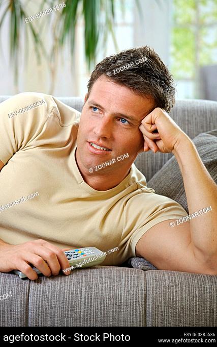 Handsome man in causal wear smiling lying on sofa with remote control handheld, watching television