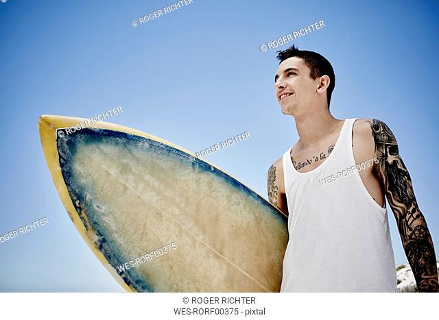 Tattooed young man with surfboard in front of blue sky