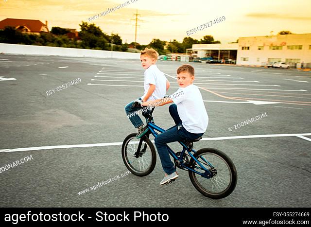 Two young brothers in white t-shirts, showing their amusing skills riding on one bike, looking into the camera. Active lifestyle concept