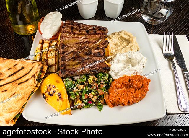 A plate of grilled beef with tabouleh salad, hummus, grilled pepper, and pita bread on a wooden table