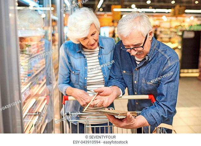 Portrait of smiling senior couple grocery shopping in supermarket, reading shopping list leaning on cart standing by freezers