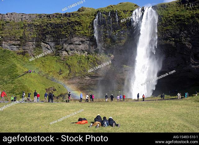 A stunning landscape of a large waterfall called Seljalandsfoss Waterfall , in Iceland, surrounded by fields and mountains, and crowds, and a rainbow