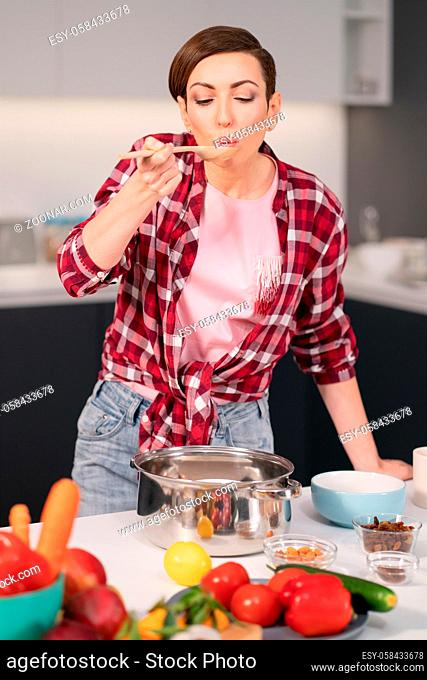 Young housewife with a short hairstyle testing a food while prepares dinner for family in the kitchen. Healthy food at home