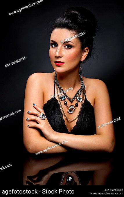 Portrait of beautiful young woman with shiny black hair and bright makeup sitting near glass table. Over dark background