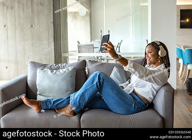 Portrait of a happy young woman listening to music with headphones and taking a selfie while leaning on a sofa at home. Concept of people in home