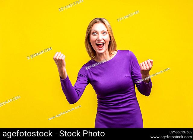 Portrait of satisfied successful woman in purple dress standing with raised fists and screaming with happiness, celebrating achievement