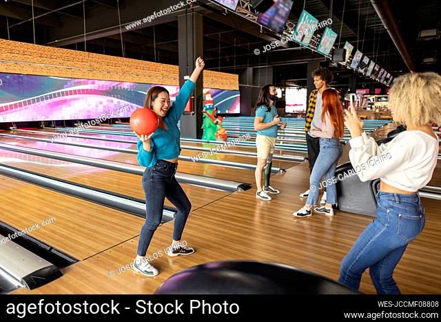 Young woman taking photo of friend holding bowling ball at alley