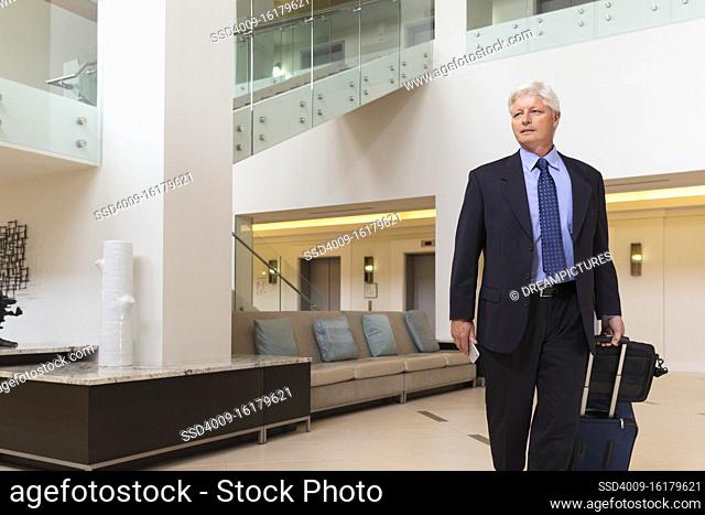 Mature Caucasian man holding cell phone walking through hotel lobby with luggage, looking off camera