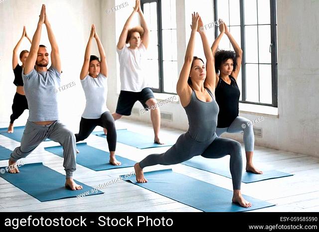 Young people practicing yoga indoor doing Warrior one exercise or Virabhadrasana 1, beautiful slim Caucasian female instructor standing in asana showing...