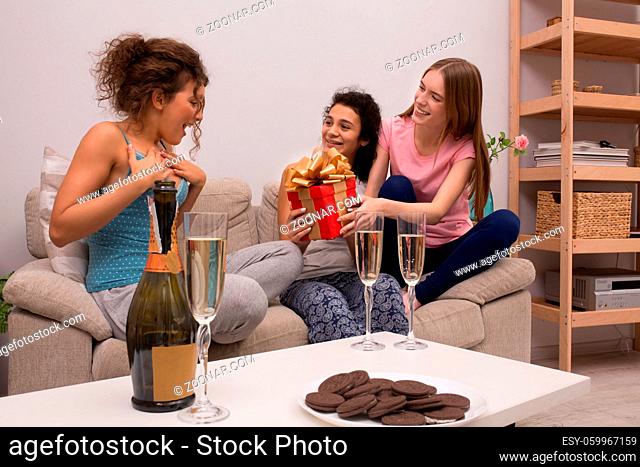 Two girls giving present to a friend. Celebrate party with champagne and cookies