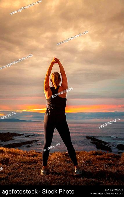 Middle-aged woman in black sportswear on her back stretching in front of the sea at sunset. Caucasian woman relaxing at dusk