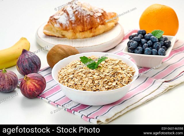 Raw oatmeal in white ceramic plate, blueberries, fig, orange, banana, on white table, breakfast