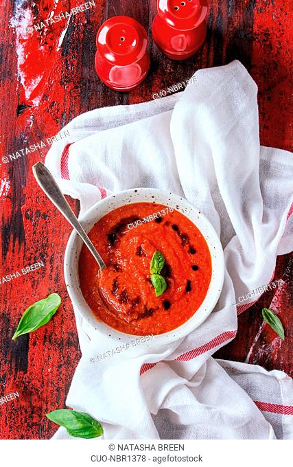 White plate of fresh cold tomato gazpacho soup with basil and balsamic, served with red salt and pepper shakers on white kitchen towel over red and black wooden...