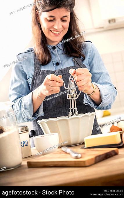 Smiling woman using egg beater while standing in kitchen at home