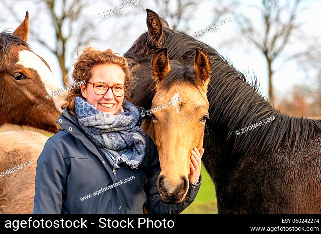 Smiling young woman in a wax coat with her 1 year old stallions in the pasture. She cuddles with them to socialize them. Three horse heads