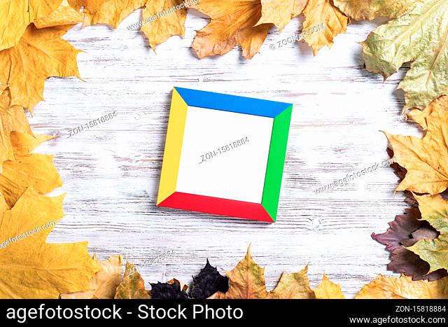 Blank rectangular photo frame lies on vintage wooden desk with bright autumn foliage. Flat lay with autumn leaves on white wooden surface