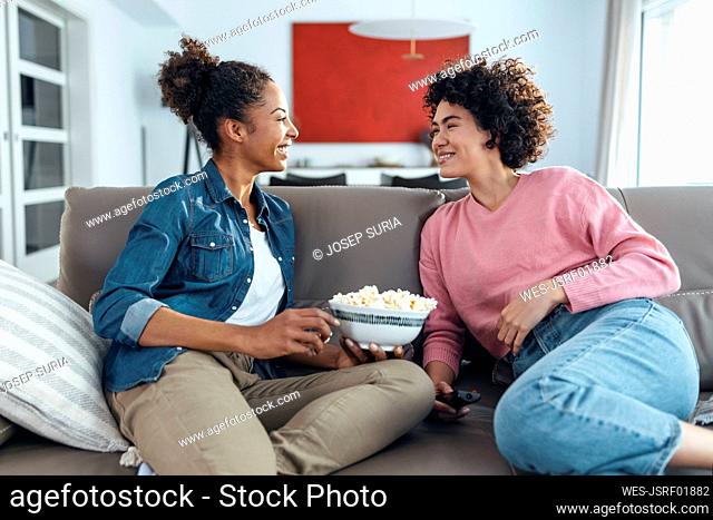 Happy woman holding bowl of popcorn talking with friend on sofa at home