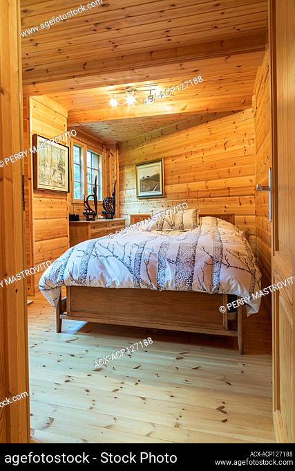 Queen size bed with wood frame and striped bedspread and matching pillows in long and narrow bedroom inside a piece sur piece Scots pine log home, Quebec