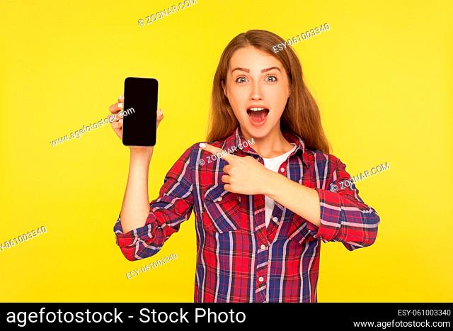 Wow, look at device. Portrait of amazed pretty girl in shirt pointing at cellphone in her hand and expressing shock surprise