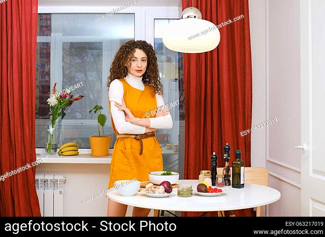 Cheerful young woman standing with crossed arms near the kitchen table with food ingredients