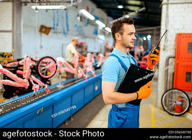 Bicycle factory, worker with a walkie-talkie poses at bike assembly line. Male mechanic in uniform installs cycle parts in workshop, industrial manufacturing