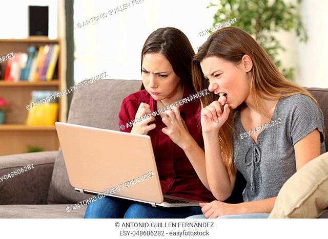 Two angry friends watching media on line with a pc sitting on a sofa in the living room at home