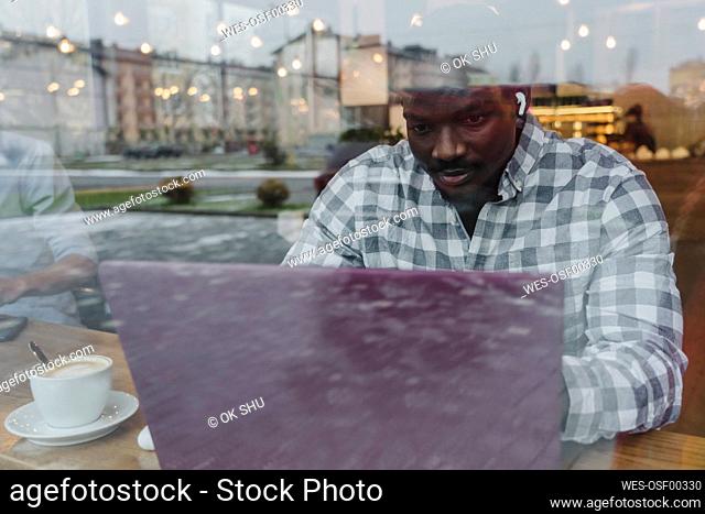 Young freelancer working on laptop seen through glass window at cafe