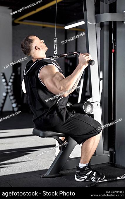 Handsome guy with huge biceps does exercise in the gym. He wears black sleeveless, shorts and sneakers. Shoot from the side. Vertical