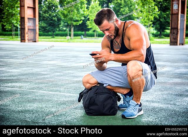 Attractive muscleman using cell phone from his bag in city park, ready for training or jogging