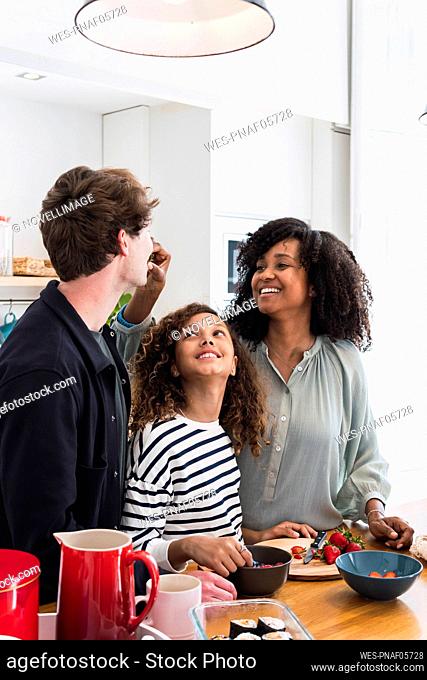cheerful family standing in kitchen mother feeding father wirh strawberry
